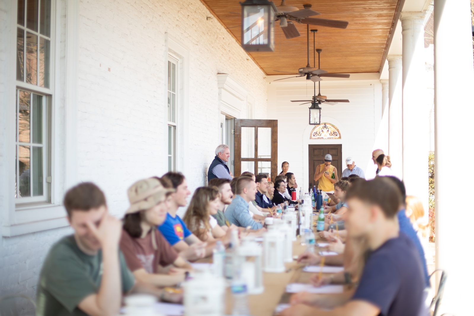 The whole group seated along one long table under a covered porch with ceiling fans.