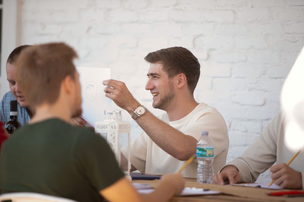 A young man smiles as he holds up his sketch to the person across the table.