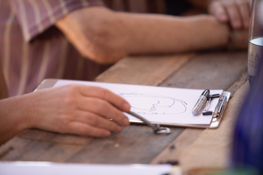 Close-up of a hand holding a pen over a sketch on a clipboard, on a weathered wooden table.