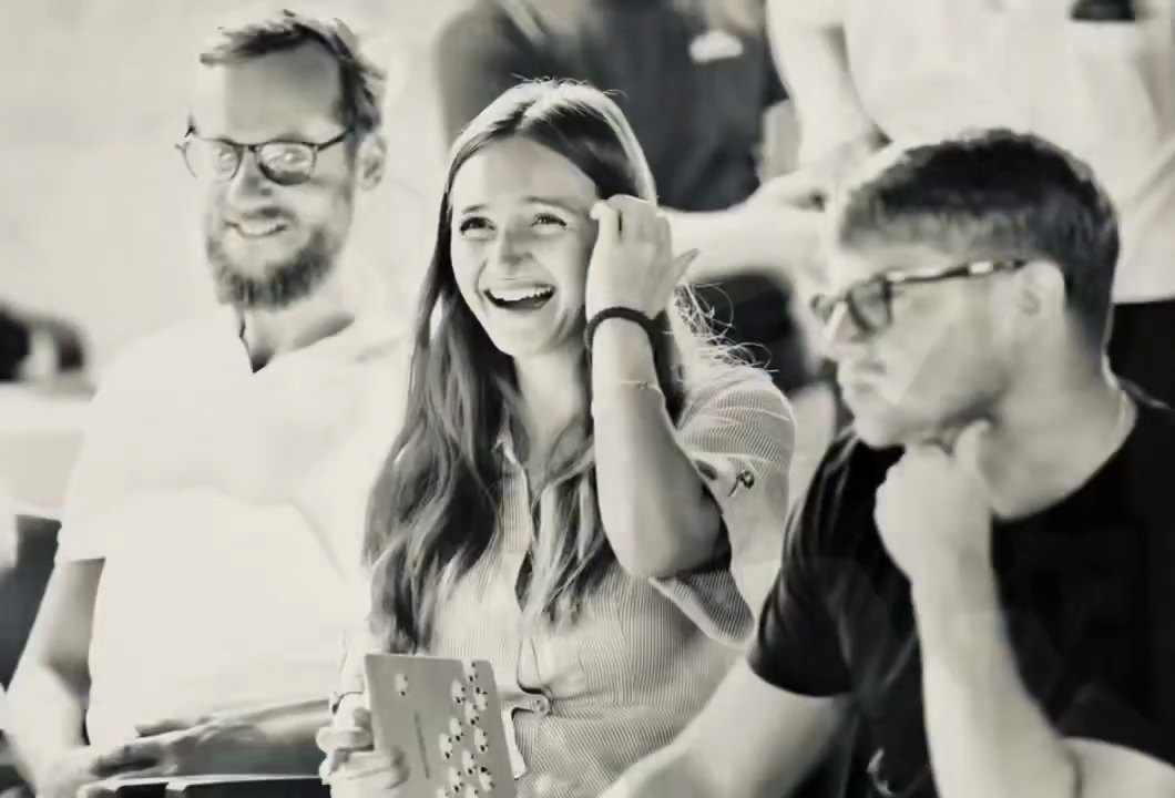 Three Builders in Milan between sessions: a young woman laughs with her hand in her hair, a bearded man in glasses smiles beside her, and a man in the foreground rests his chin on his hand.